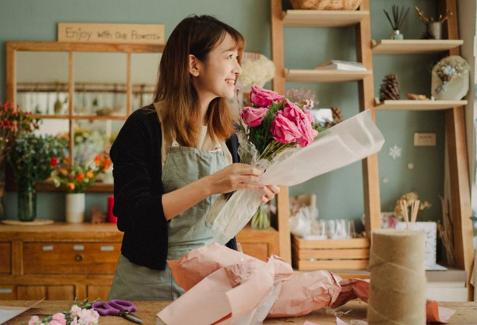 A young woman with shoulder-length brown hair is in a flower shop, smiling as she holds a bouquet of fresh pink roses, lilies, and smaller pink flowers wrapped in white paper. She is wearing a light-c