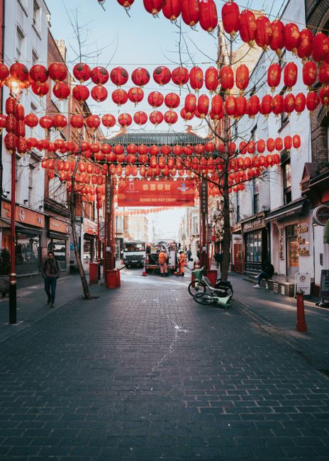 The image depicts a lively street scene in Chinatown, decorated with numerous bright red Chinese lanterns hanging in rows across the street. The lanterns are round, made of paper, and vibrant in color