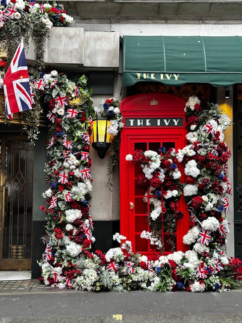 A vibrant floral arrangement surrounding a red British telephone booth, decorated with white, red, and navy blue flowers, including carnations and chrysanthemums, accented with Union Jack flags. The f