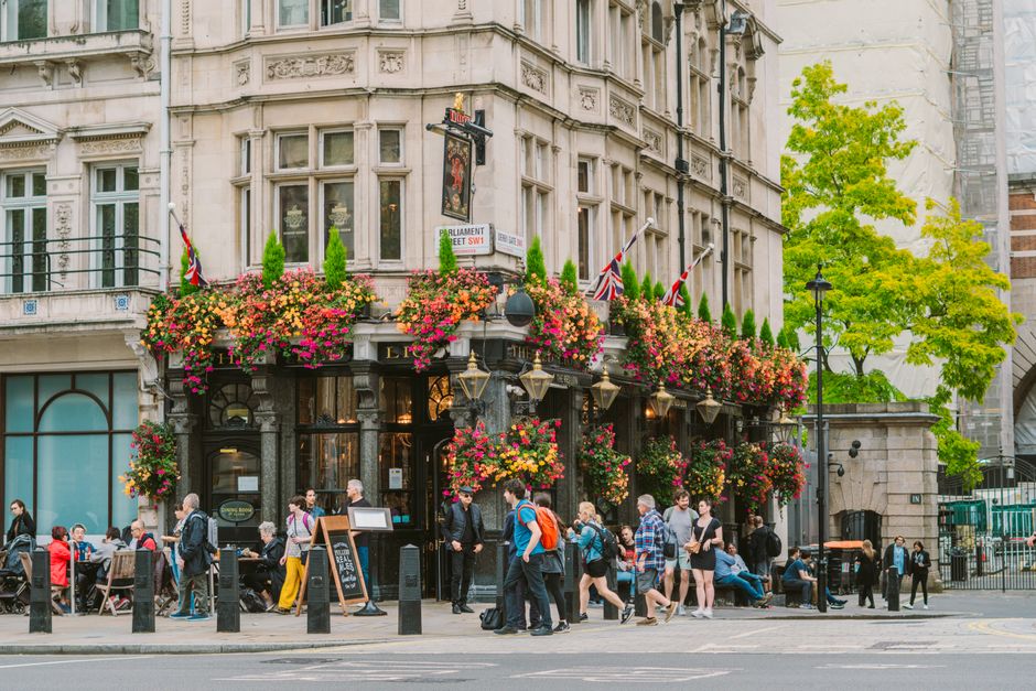 A lively street scene outside a traditional English pub in Leicester Square, decorated with abundant floral arrangements of pink, orange, and purple flowers cascading from hanging baskets and window b