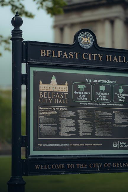 Close-up of a dark green Belfast City Hall signboard with gold lettering and emblem at the top, displaying visitor attractions, guided tours, self-guided visitor exhibitions, and the Bobbin Coffee Sho